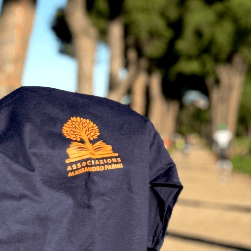 Maglietta blu con il logo arancione dell'Associazione Alessandro Parini, appesa tra gli alberi di un parco.