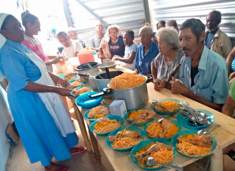 Una suora serve piatti di riso e verdure a una fila di persone anziane sedute a un tavolo, in una mensa popolare a Cuba.