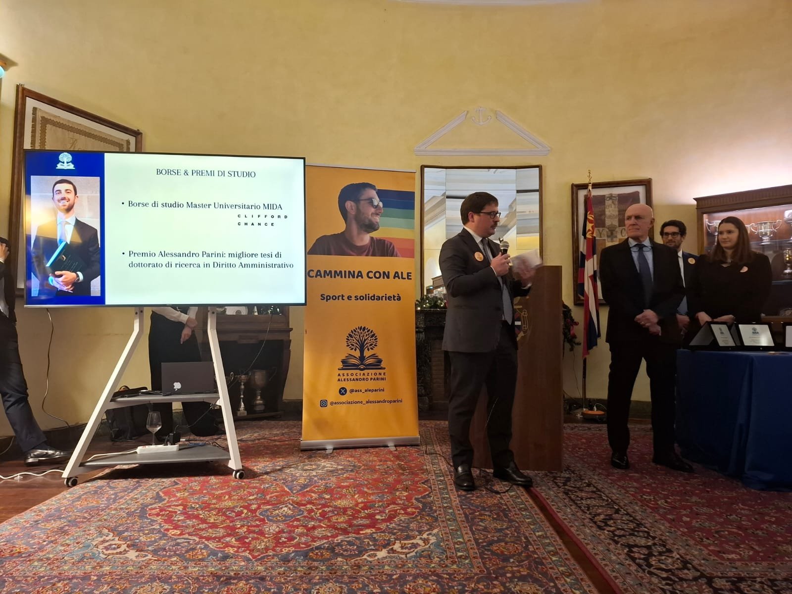 Un uomo parla al leggio accanto a uno schermo con la scritta «Borse & premi di studio» e al banner della Cammina con Ale.