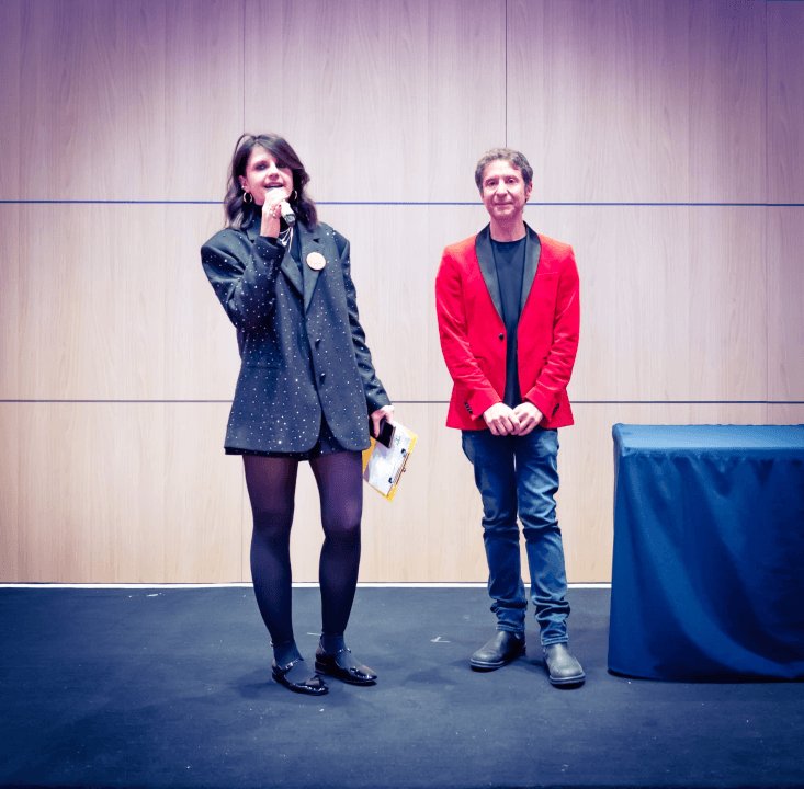 Sul palco, una donna parla al microfono accanto a un ragazzo in giacca rossa.